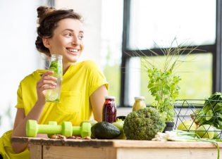 Portrait of a young sports woman in yellow t-shirt sitting indoors with healthy food and dumbbells on the table